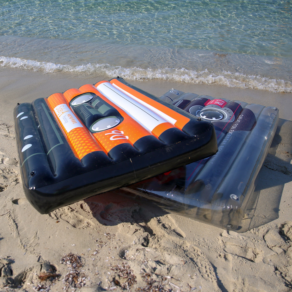 Two cassette pool floats on the edge of the shore. The black cassette is angled on top of the clear cassette pool float. 