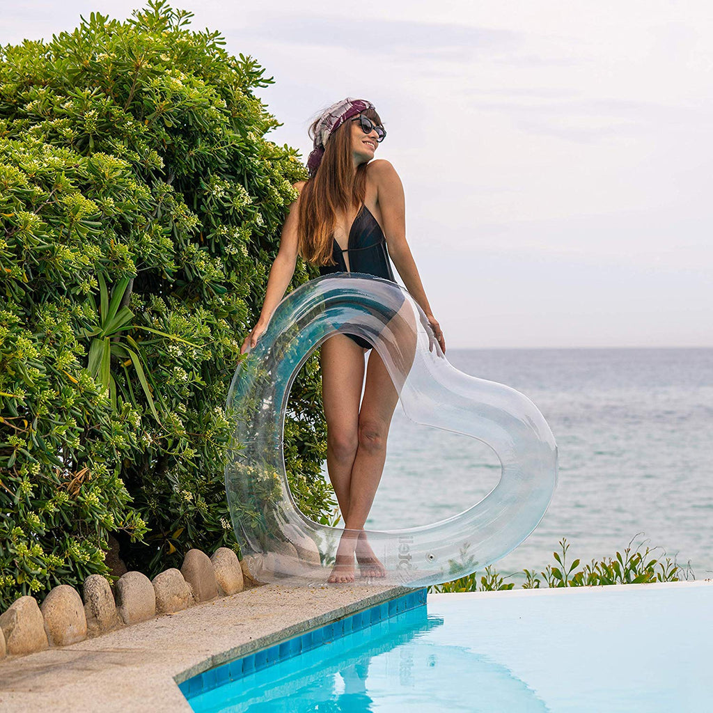Woman in black swimsuit poses with legs crossed behind clear heart float from the best pool float brand. She's standing next to an infinity pool in front of lush plants