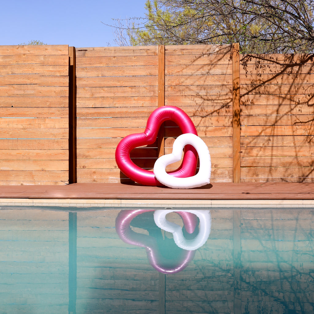 the metallic pink heart shaped pool float and the mini white heart shaped pool float resting on top of each other in front of a fence by the pool