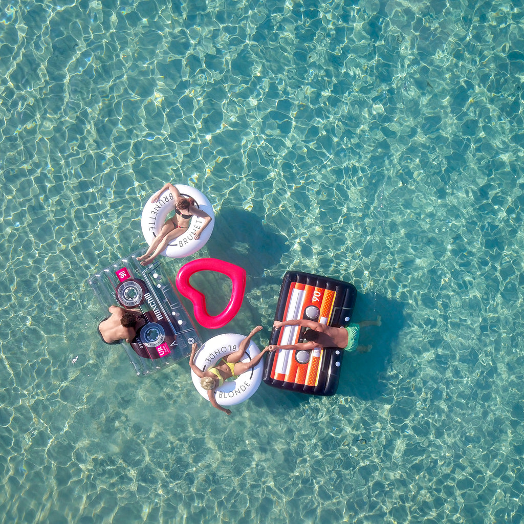 Aerial photo of two couples floating near to one another on top of five floats. Woman on left is sitting in a white swim ring with the word brunette on it. Her feet are resting on a clear cassette float that a man is leaning over onto from the water. The man on the right is reaching over a black cassette float from the water and holding onto the foot of a woman on a white swim ring with the word blonde on it. Between the two couples playing in the water there is a red heart shaped pool float. 