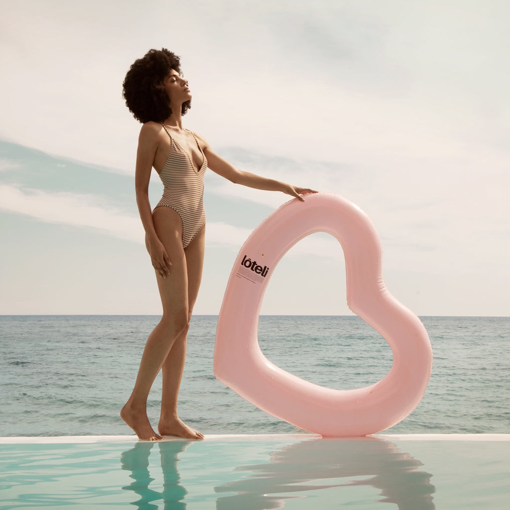 Girl in striped bathing suit holding pink heart shaped pool float at border of pool with sea in the background