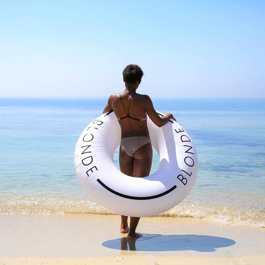 Woman in swimwear facing away, standing on a beach holding a white inflatable innertube around her torso.
