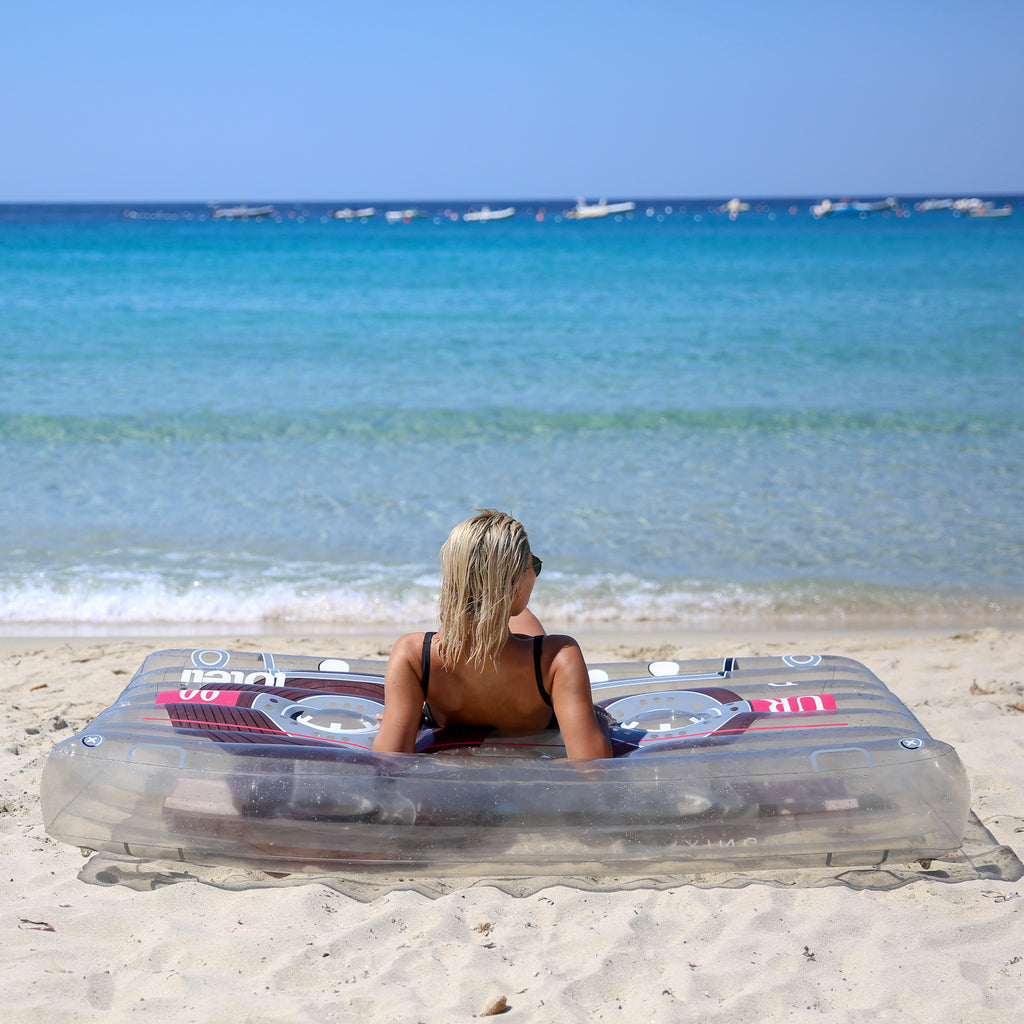 Woman lying on her back propped up on her elbows on top of a clear cassette pool float staring out at the ocean. 
