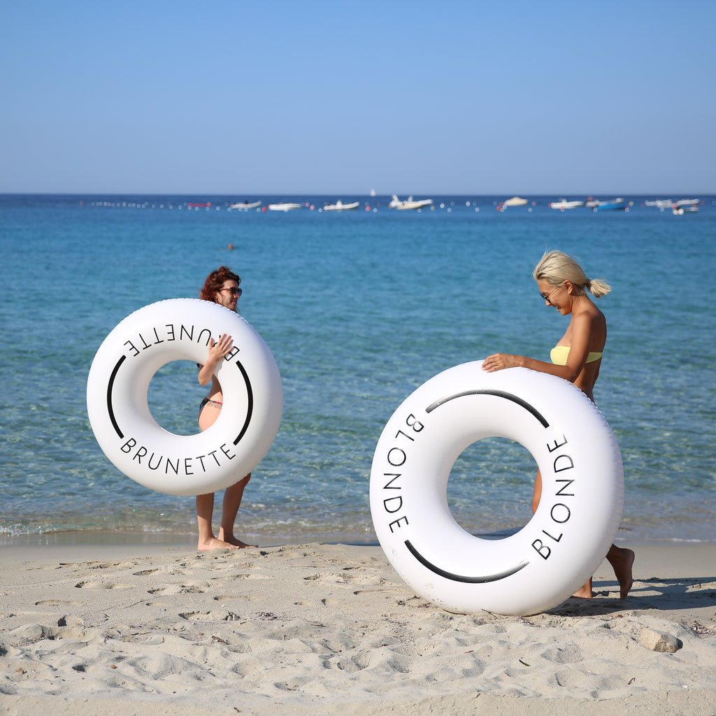 Two women standing near the sea shore, woman on left is holding a white swim ring with the word brunette on it with her right arm. The woman on the right is laughing and leaning on a white swim ring with the word blonde on it. 