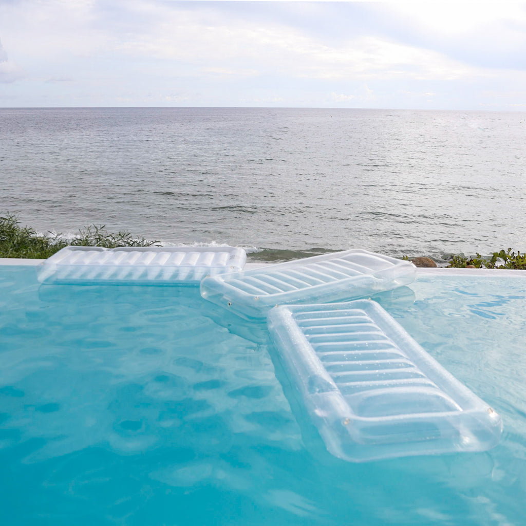 Three clear lounger floats from the best pool float brand are at the edge of a pool that overlooks the sea