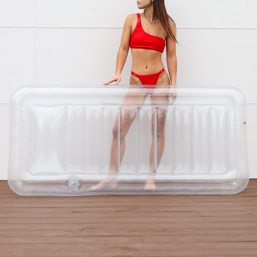 Woman in red swimsuit posing behind the clear lounger pool float. She is looking to the left and part of her head is obstructed.