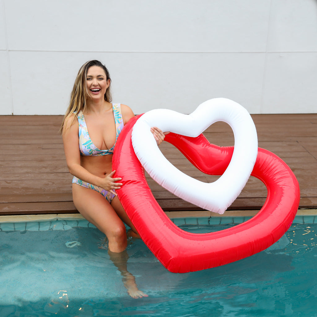 Woman posing with red heart shaped pool float and its perfect pair, the mini white heart pool float.  The mini fits inside the full-size for a great instagram photo
