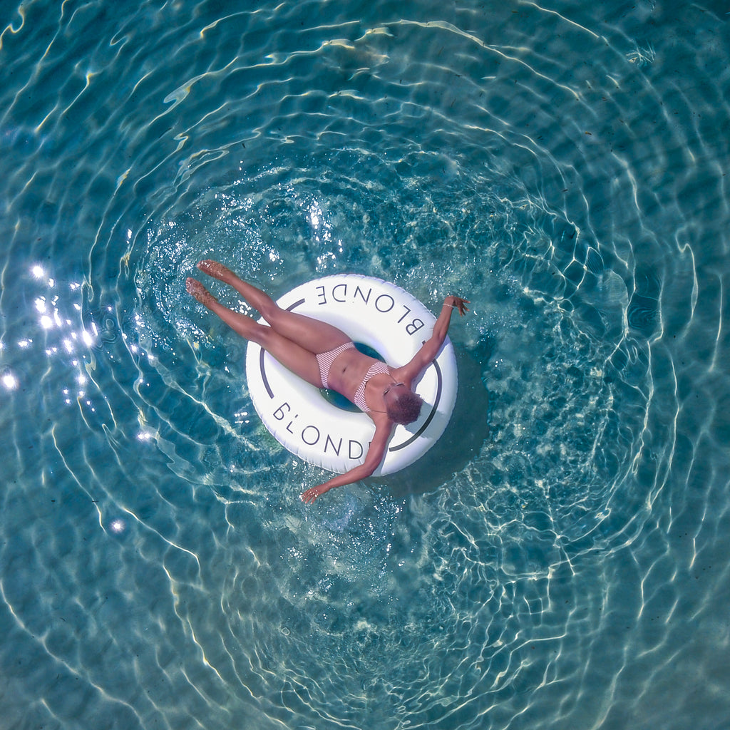 Aerial photo of woman laying in a white inflatable swim ring with the word Blonde on it in the center of a pool. 