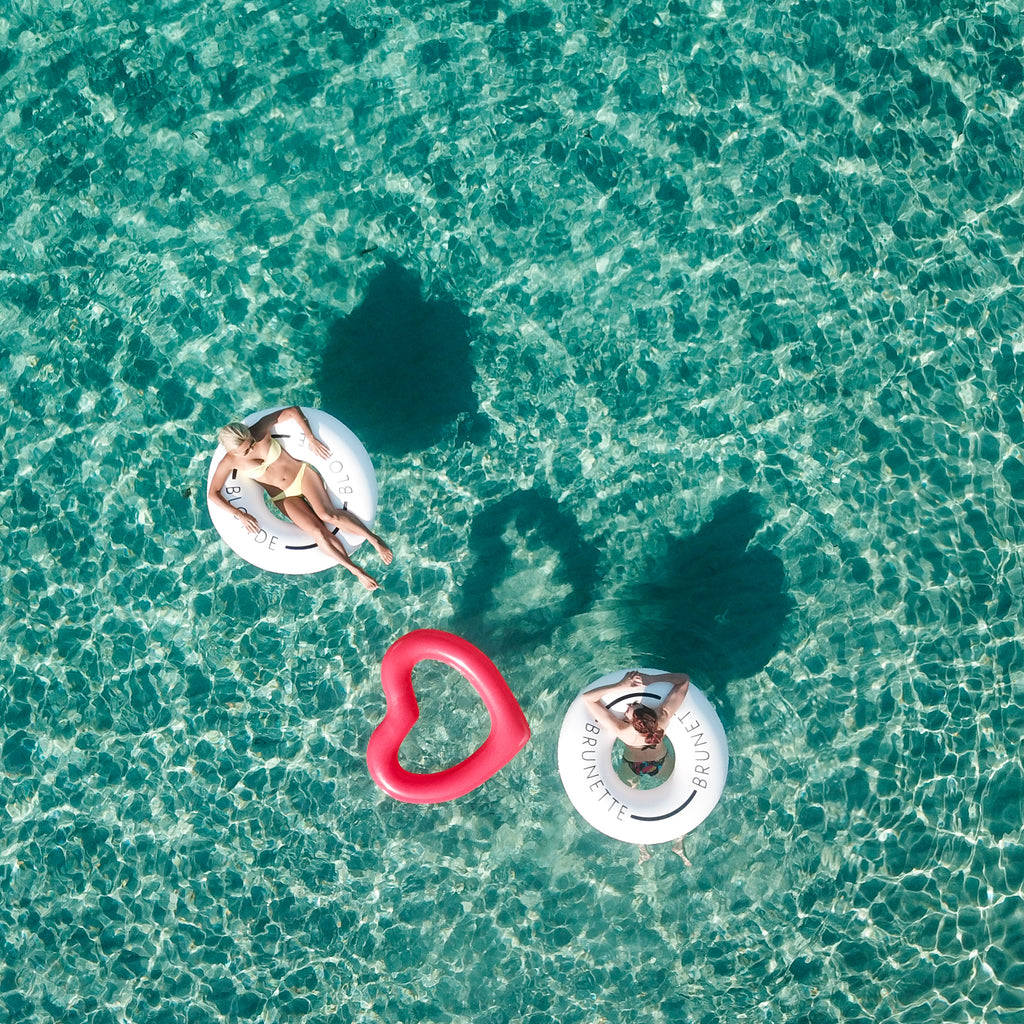 Aerial photo of two women in swim rings floating in the sea. The woman on the left in on a swim ring with the word blonde on it. The woman on the right in floating in a white swim ring that says brunette. There is a red heart shaped swim ring floating between the two. 