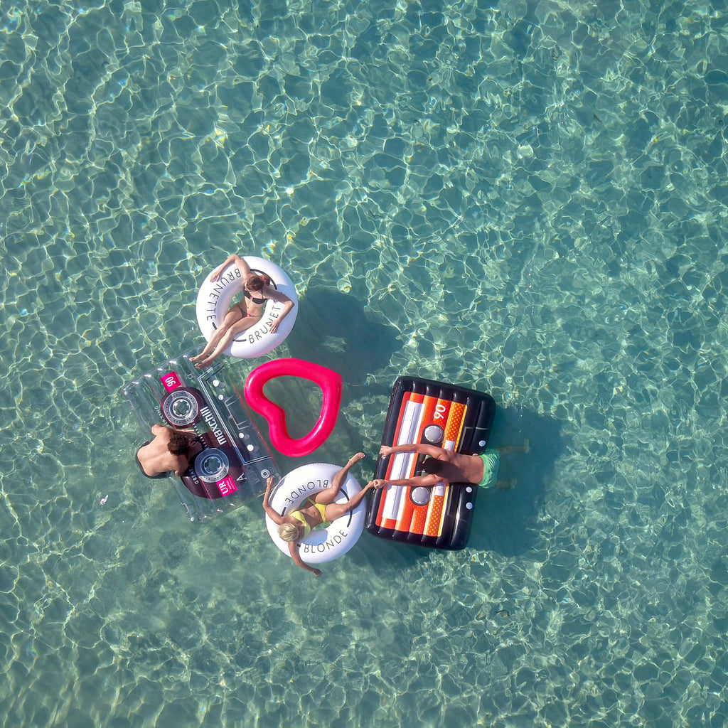 Ariel photo of an assortment of floats being split by friends in the sea.  There are two women on the brunette and blonde floats, a red heart float between them, a man on a clear cassette float to the left and a man on a mixtape pool float to the right