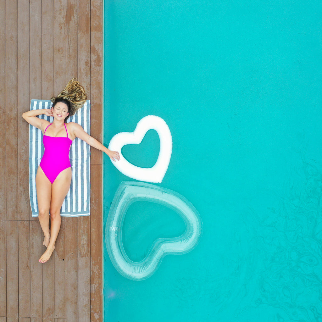 Woman in purple lays beside pool, arm outstretched to the mini white heart float, with an adult size clear heart float below it