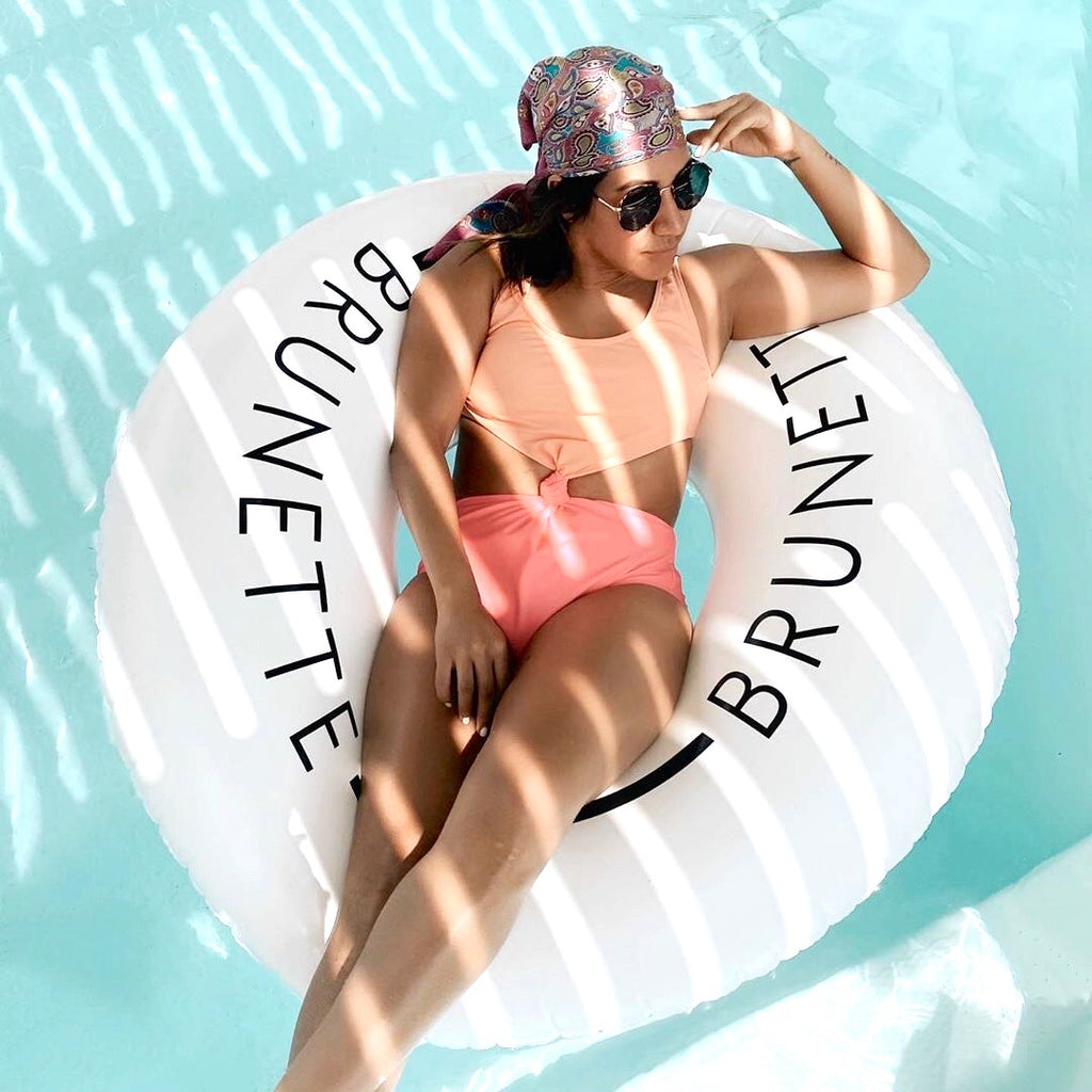 Woman in peach and pink swimwear wearing a colorful paisley patterned bandana on her head and black aviator sunglasses is lounging back on a white swim ring with the word brunette on it. The woman is staring into the distance.