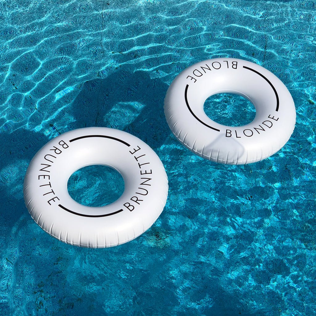 Two white pool float rings, one with the word Brunette, on with the word Blonde on it, floating in a pool.