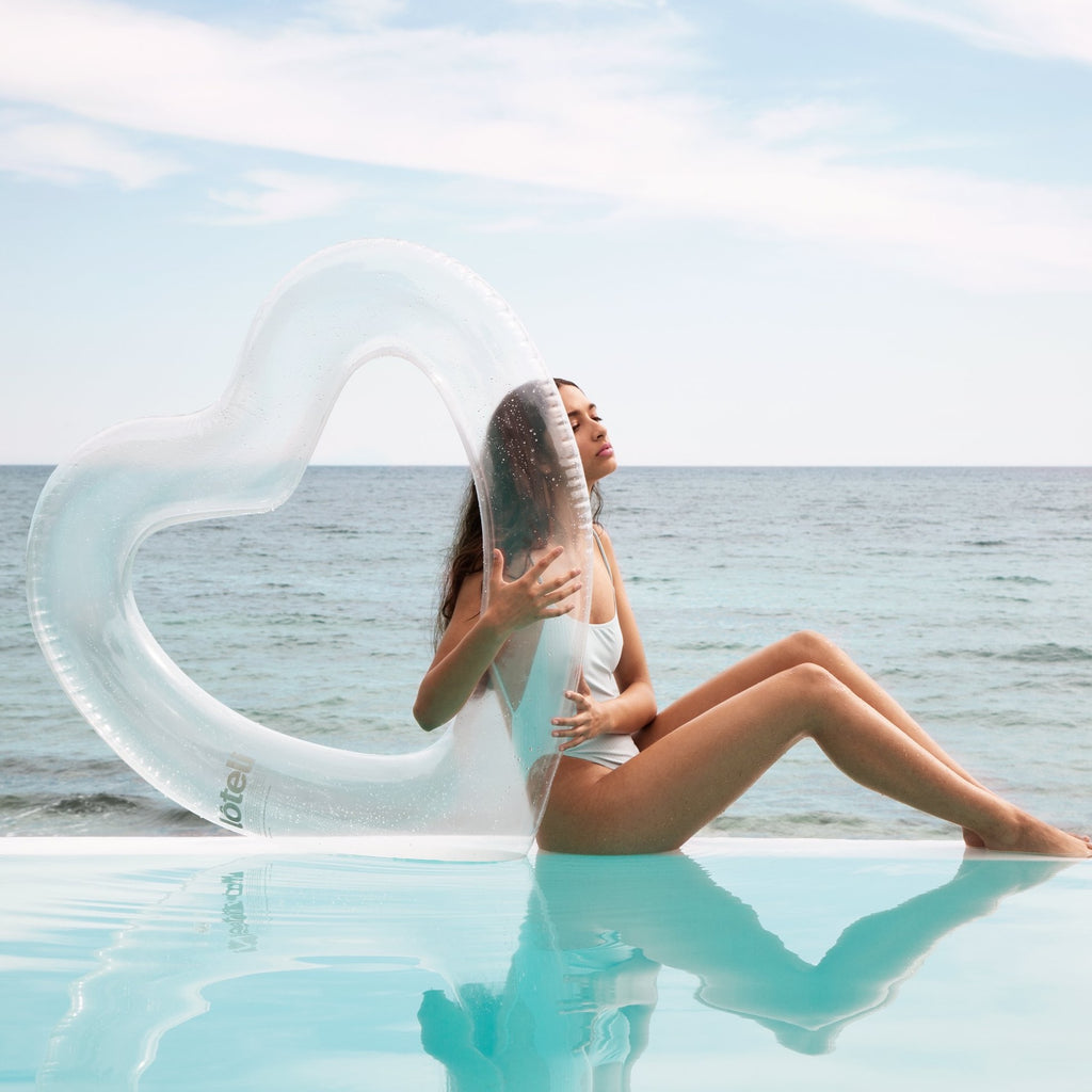 Woman in white bikini is sitting on the edge of an infinity pool, embracing her clear heart float. Her eyes are closed