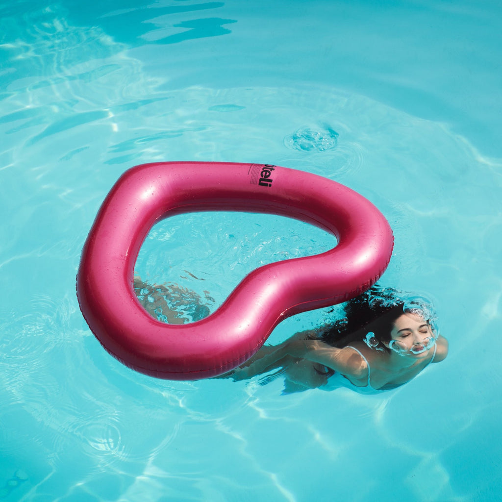 woman in white bathing suit breaks surface of pool in front of metallic pink heart pool float