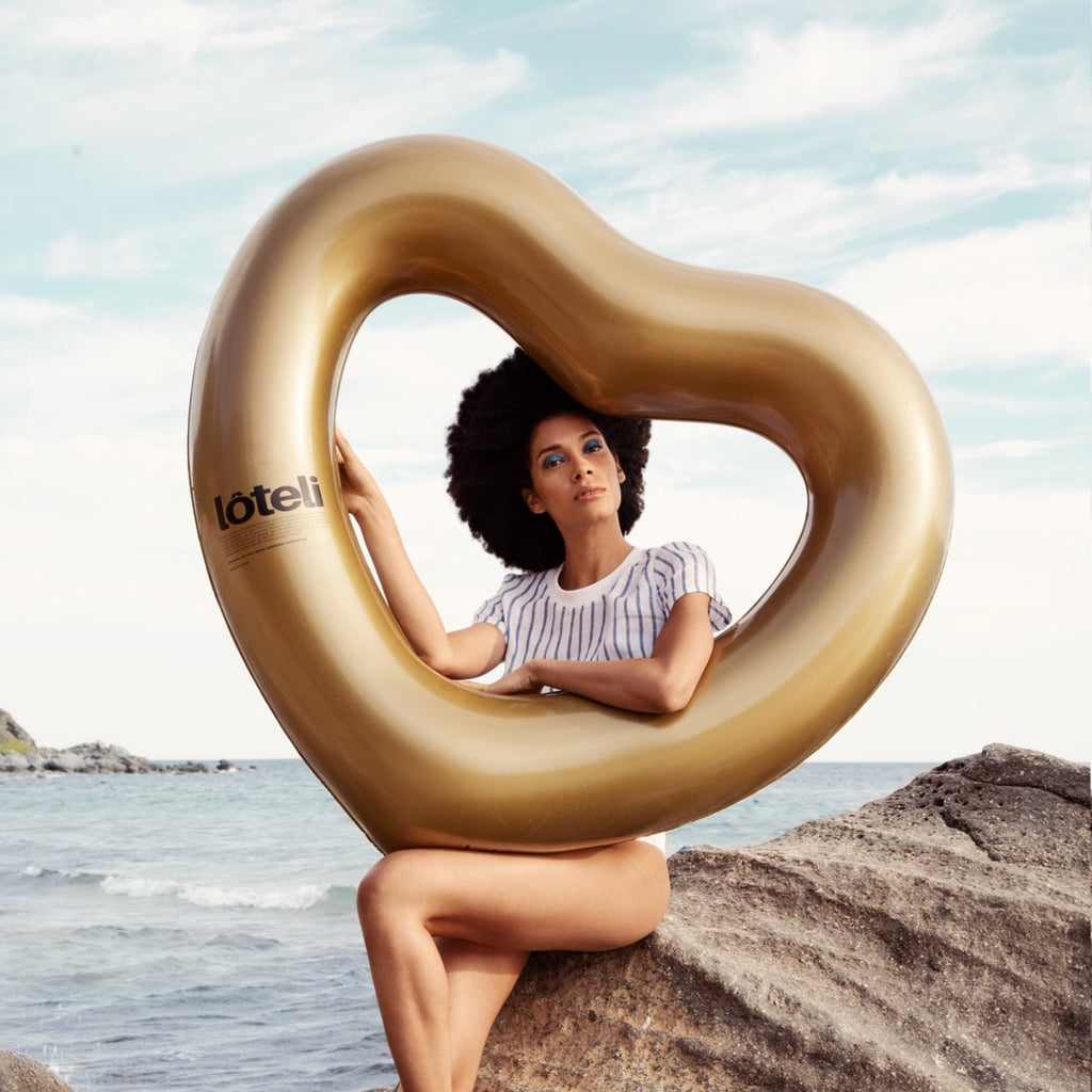 Woman posing on rocks by the beach with gold heart float resting on her leg. She is looking directly into the camera