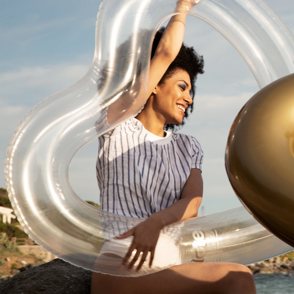 Woman in white swimsuit holding her clear heart float, ready to dive into the water. In front of her is a gold heart float from lôteli