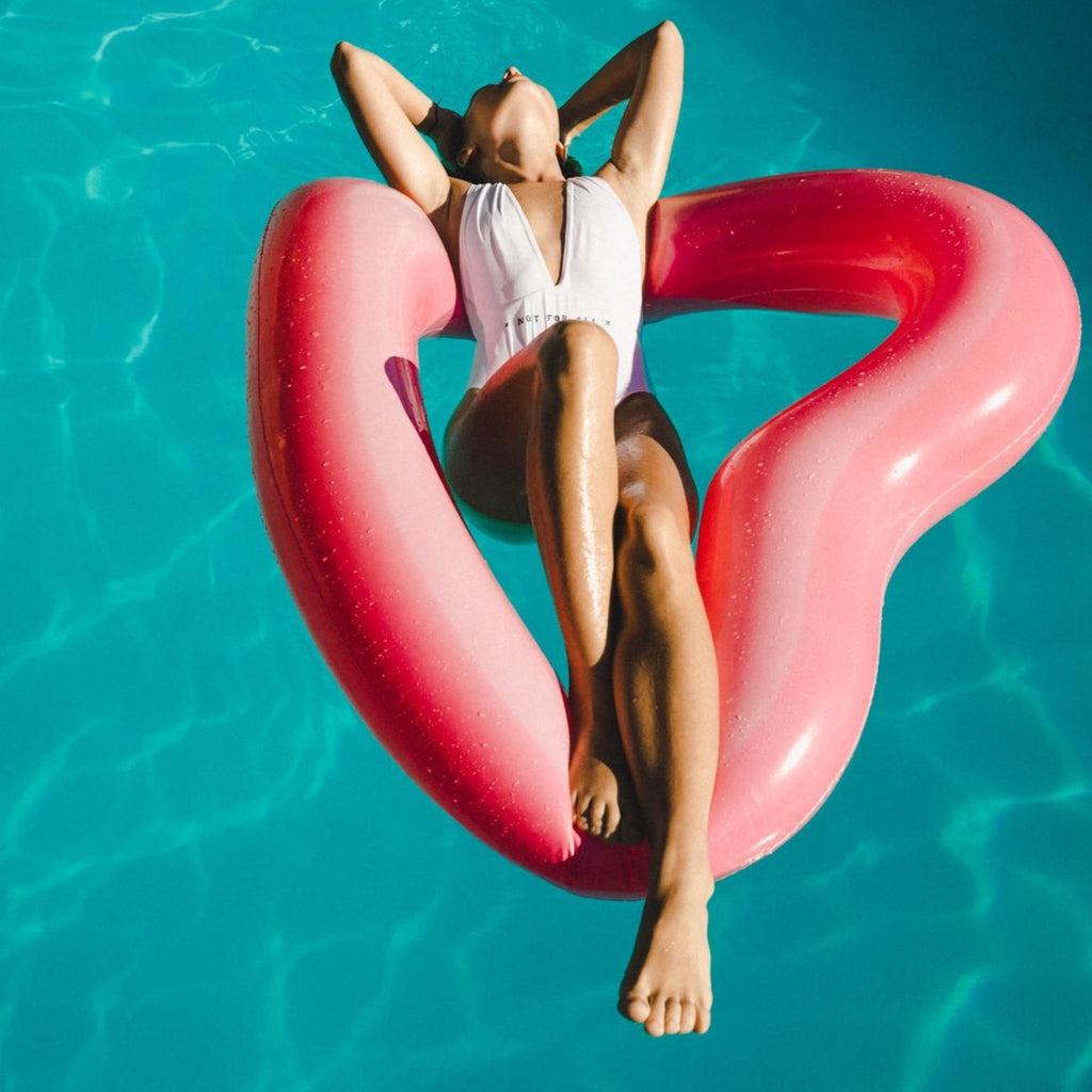 Girl in white swimsuit leaning back with hands on head while floating in the pool on a red heart shaped float